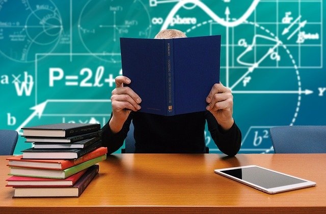 Nauczyciel trzymający otwartą książkę, zasłaniającą całą twarz, siedzący przy biurku na którym leży po lewej stronie w słupku kilka książek z kolorowymi okładkami, a po prawej tablet. Z tyłu za nauczycielem tablica z wzorami matematycznymi