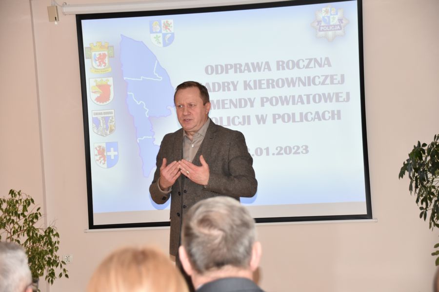Mężczyzna w brązowym garniturze, Starosta Policki na tle slaidera prezentacji z odprawy policyjnej.