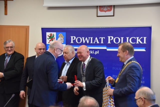 Obrady sesji Rady Powiatu w Policach, na tle grafiki niebieskiej z dużym herbem Powiatu Polickiego stoi sześciu mężczyzn, następuje wręczenie medalu i gratulacji od Przewodniczącego Rady Powiatu w Policach i Starosty Polickiego
