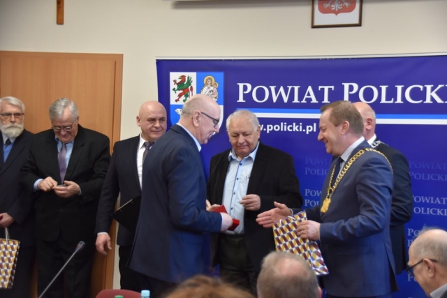 Obrady sesji Rady Powiatu w Policach, na tle grafiki niebieskiej z dużym herbem Powiatu Polickiego stoi siedmiu mężczyzn, następuje wręczenie medalu i gratulacji od Przewodniczącego Rady Powiatu w Policach i Starosty Polickiego