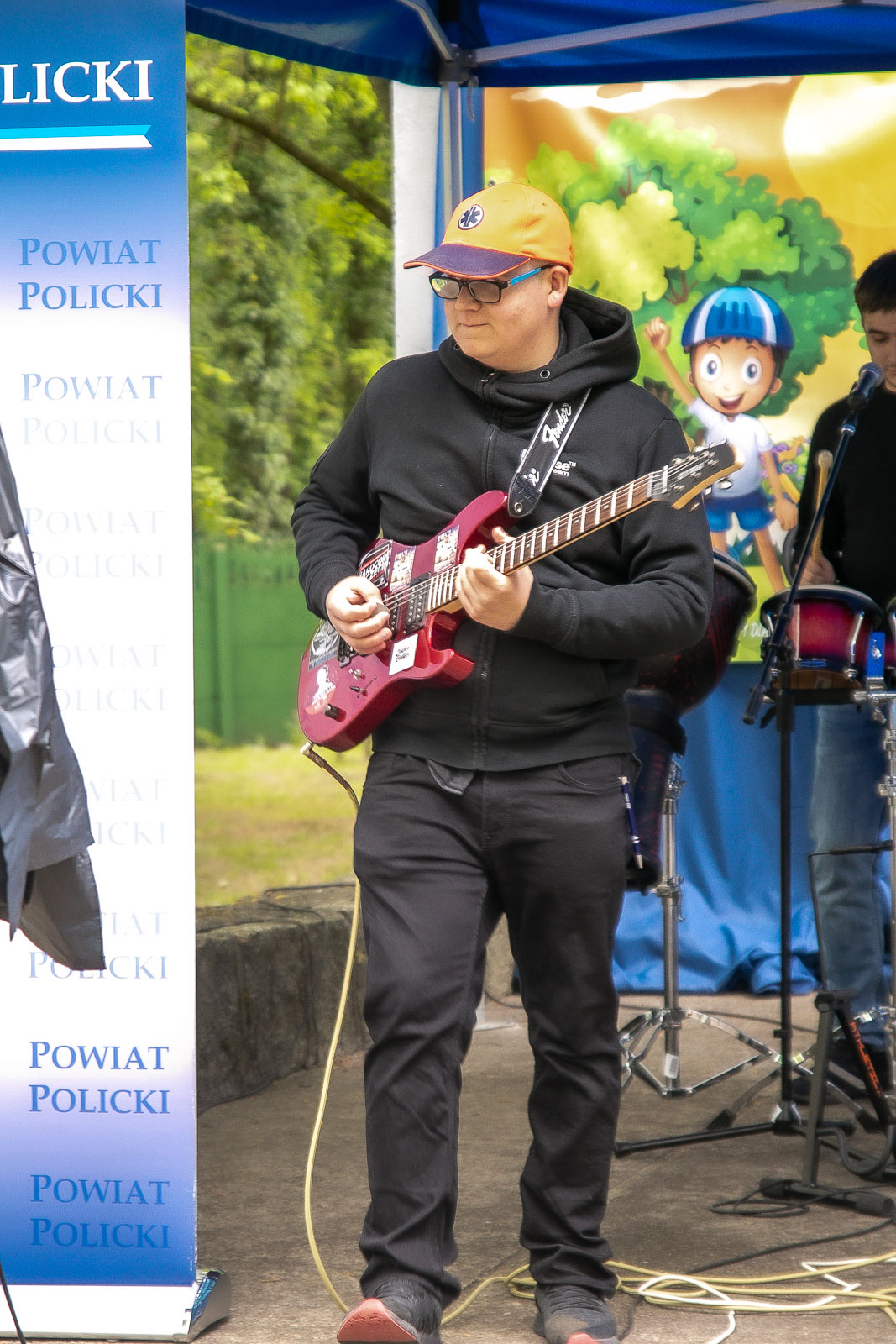 Mężczyzna gra na czerwonej gitarze elektrycznej, w tle widoczna jest ścianka Powiatu Polickiego