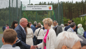 Tłum ludzi zebrany przy wejściu na kompleks boisk sportowych .W tle tablica z napisem kompleks boisk sportowych im. Jana Matury.