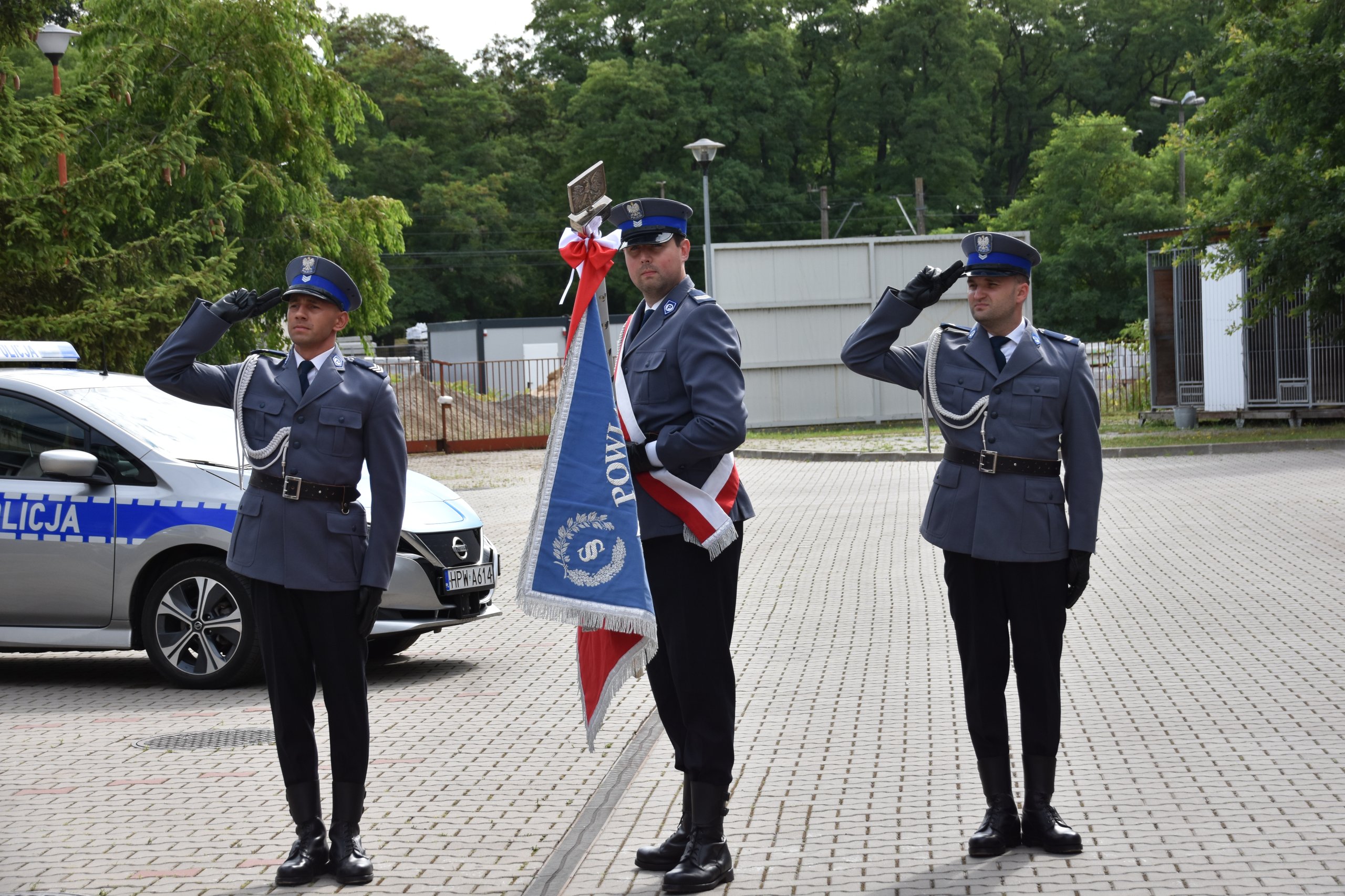Trzech mężczyzn, funkcjonaiuszy Komedny Powiatowej Policji w Policach w poczcie sztandarowym na dziedzińcu komendy.