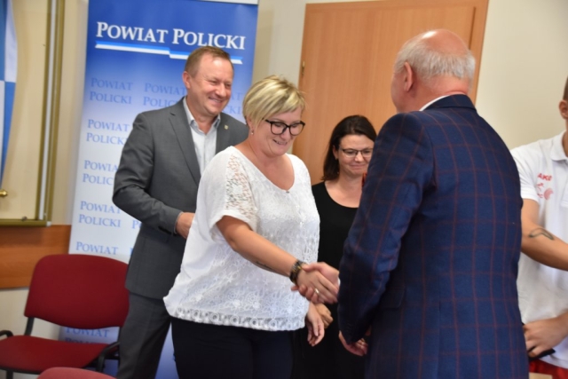 Moment składania gratulacji dla mamy Oliwii Styczyszyn przez Przewodniczącego Rady Powiatu w Policach. Mężczyzna podaje rękę kobiecie, w tle pozostali uczestnicy wydarzenia.i