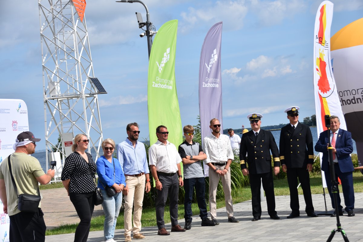 Mieszana grupa ludzi, mężczyźni, kobiety, przedstawiciele organizatorów 59. Etapowych Regat Turystycznych, stojący na dziedzińcu Mariny Trzebież w słoneczny dzień podczas oficjalnego podsumowania zawodów.