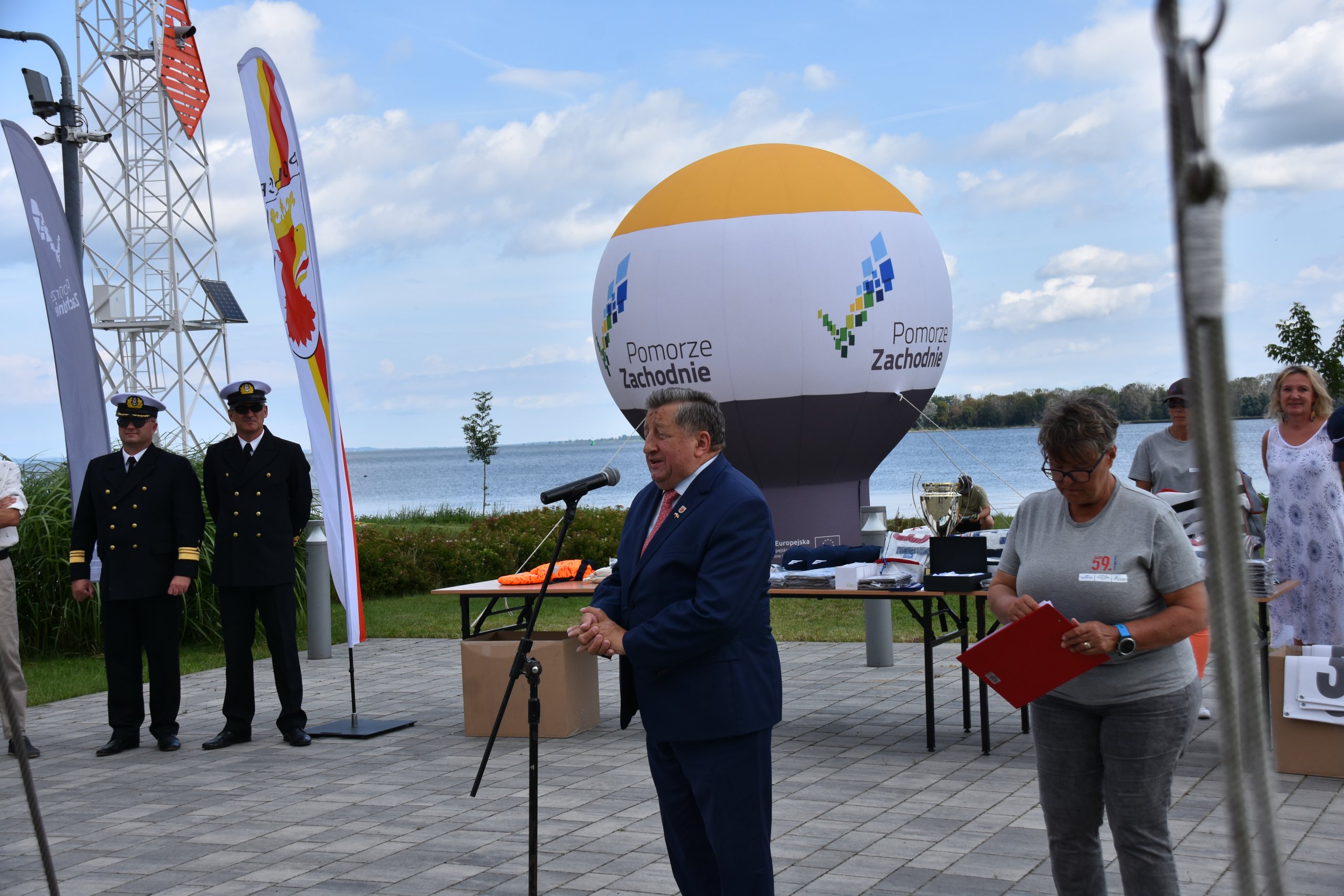 Władysław Diakun Burmistrz Polic przemawiający przez mikrofon do wszystich uczestników regat, w tle widoczny jest balon z logiem pomorza zachodniego.
