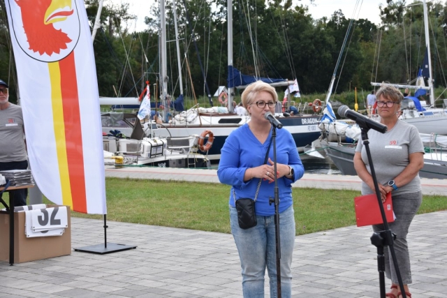 Wicestarosta Joanna Napiwodzka składająca przez mikrofon serdeczne gratulacje dla wszystkich uczestników 59. Etapowych Regat Turystycznych, w tle widoczne są łódki na wodzie