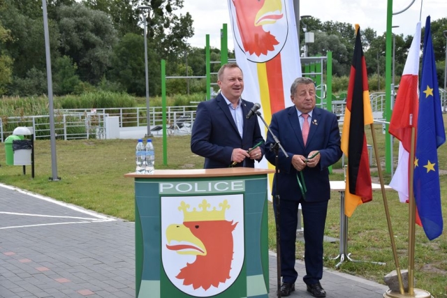 Przy mównicy z grafiką gminy Police stoi w ciemnym garniturze męźczyzna Starosta Policki, przemawiający do mikrofonu, obok stoi drugi mężczyzna w ciemnym garniturze, Burmistrz Polic w tle zielona trawa i stojak z flagami Niemiec, Polski i Unii Europejskiej