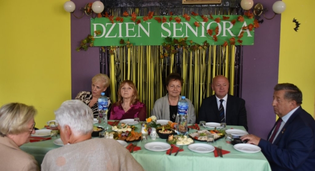 Przy stole zakrytym zielonym obrusem, zasiada pięć kobiet i dwóch mężczyzn, a nad nimi na przyozdobionej ścianie powieszony zielony plakat z białym napisem Dzień Seniora