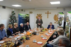 W tle mężczyzna przemawiający, pozostałe osoby siedzą przy stole