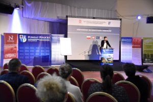 W tle stoi kobieta przy mównicy