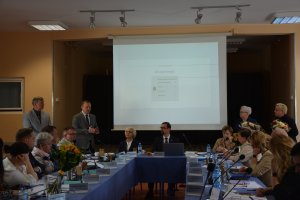 W tle telebim, 2 stojące osoby i siedzące osoby przy stole