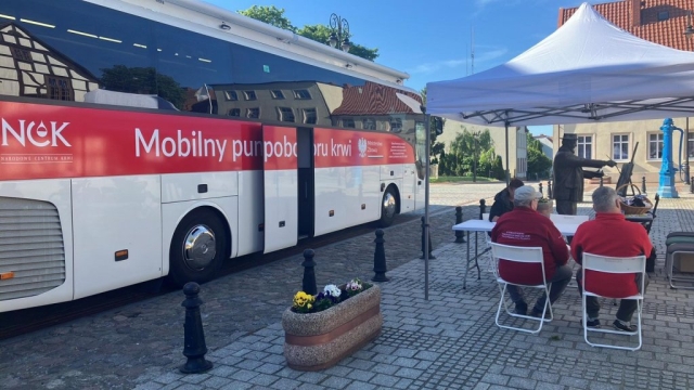 Autobus Mobilnego punktu poboru krwi stojący przy budynku, w tle zabudowania mieszkalne, a także osoby siedzące na krzesełkach przy stole, oczekujące do oddania krwi