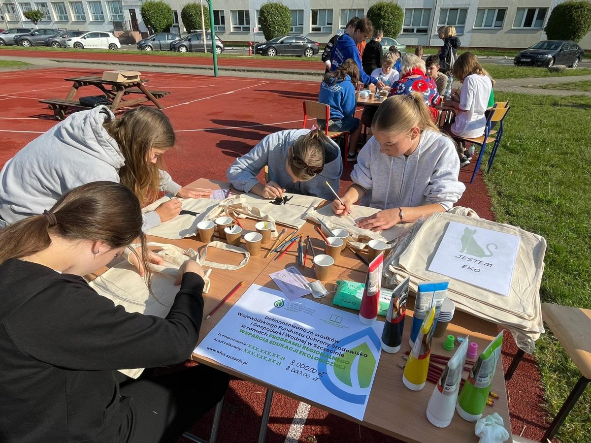 Grupa młodzieży przygotowuje ekologiczne torby przy stole wystawionym na szkolnym boisku.