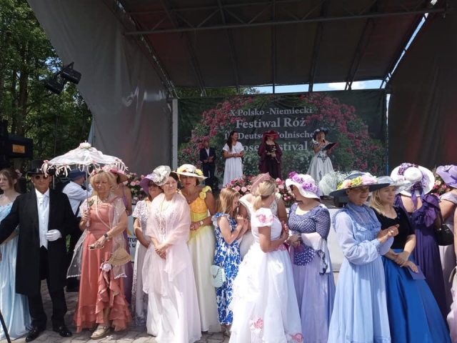 Kobiety w kolorowych sukienkach i kapeluszach stojące przed sceną w plenerze