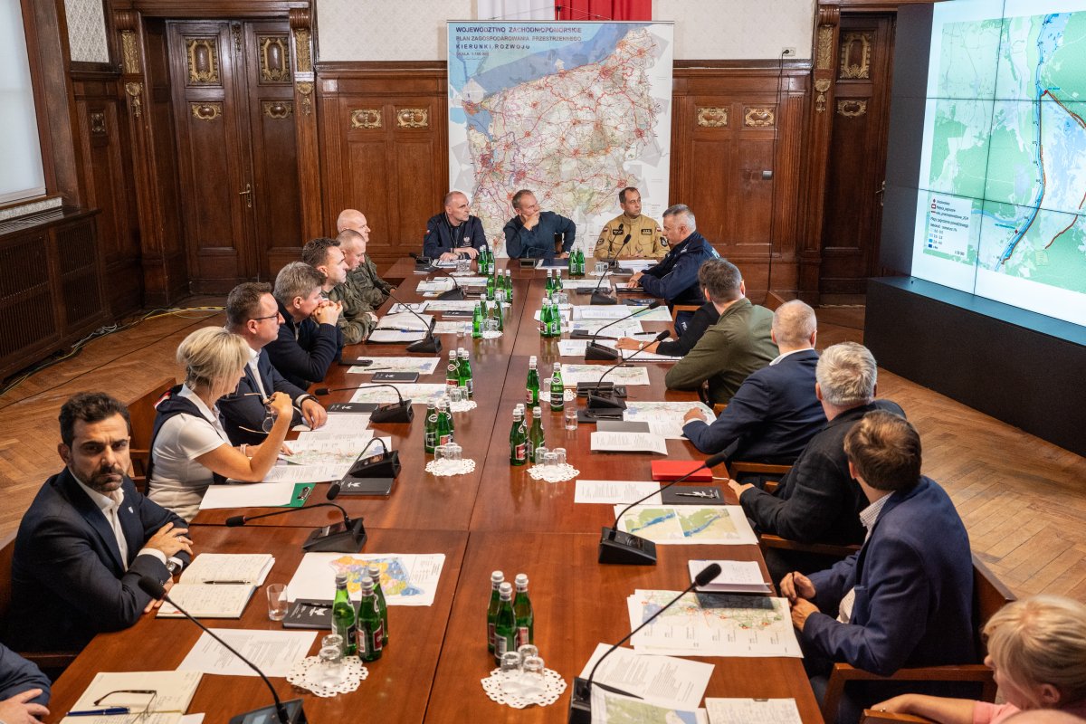 Uczestnicy posiedzenia siedzący przy długim stole po obu stronach, w tle ekrany led, mapy województwa.