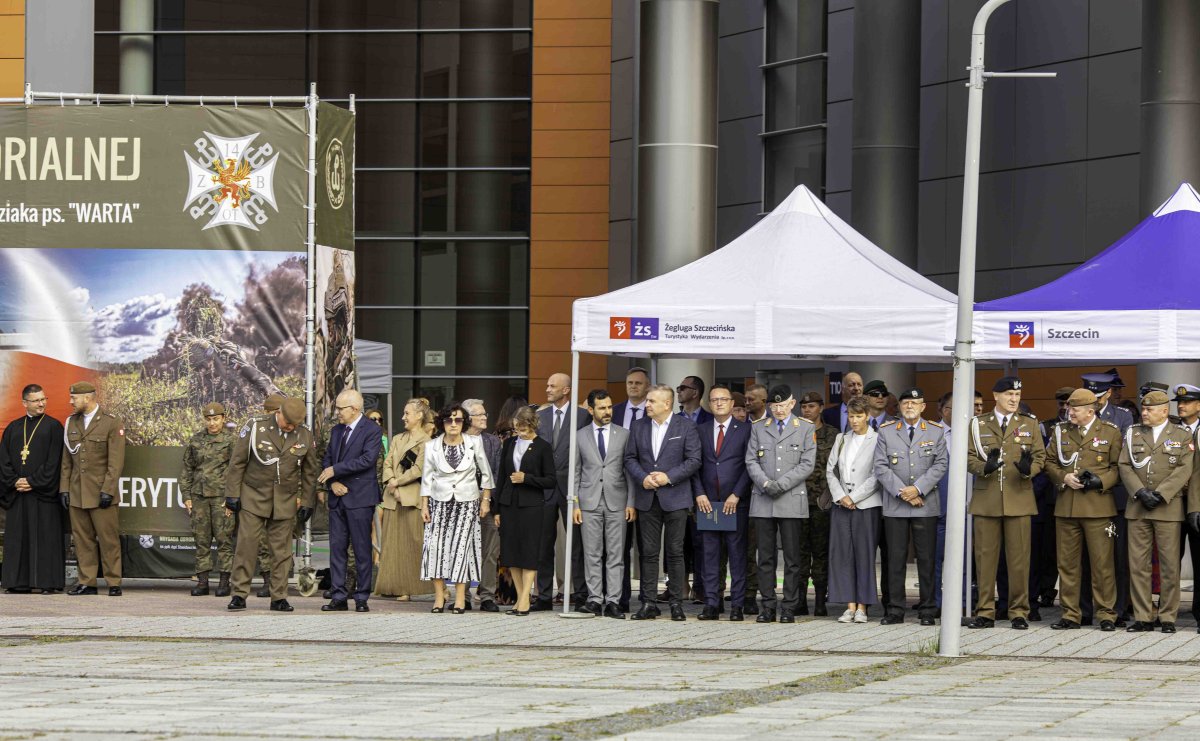 Grupa uczestników apelu z wojskowym ceremoniałem stojąca pod namiotem na placu przed halą sportową.