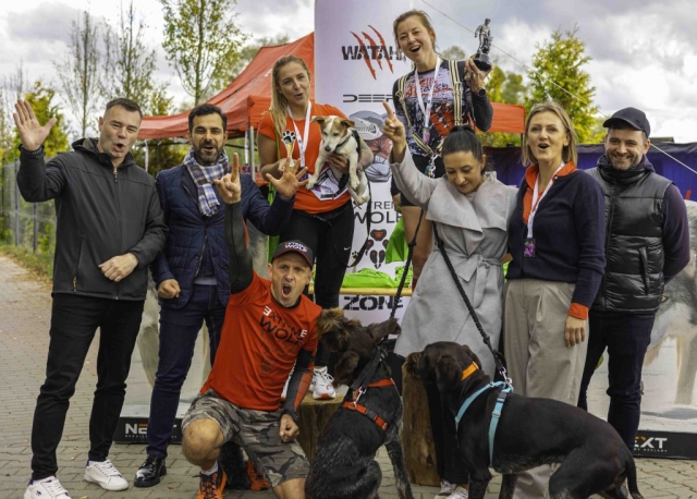 Grupowe zdjęcie osób pozujących z uśmiechem, trzymających psy ze schroniska. Każdy pies jest innej rasy i wielkości, a wszyscy wyglądają na zadowolonych. Tło zdjęcia to otwarta przestrzeń na świeżym powietrzu